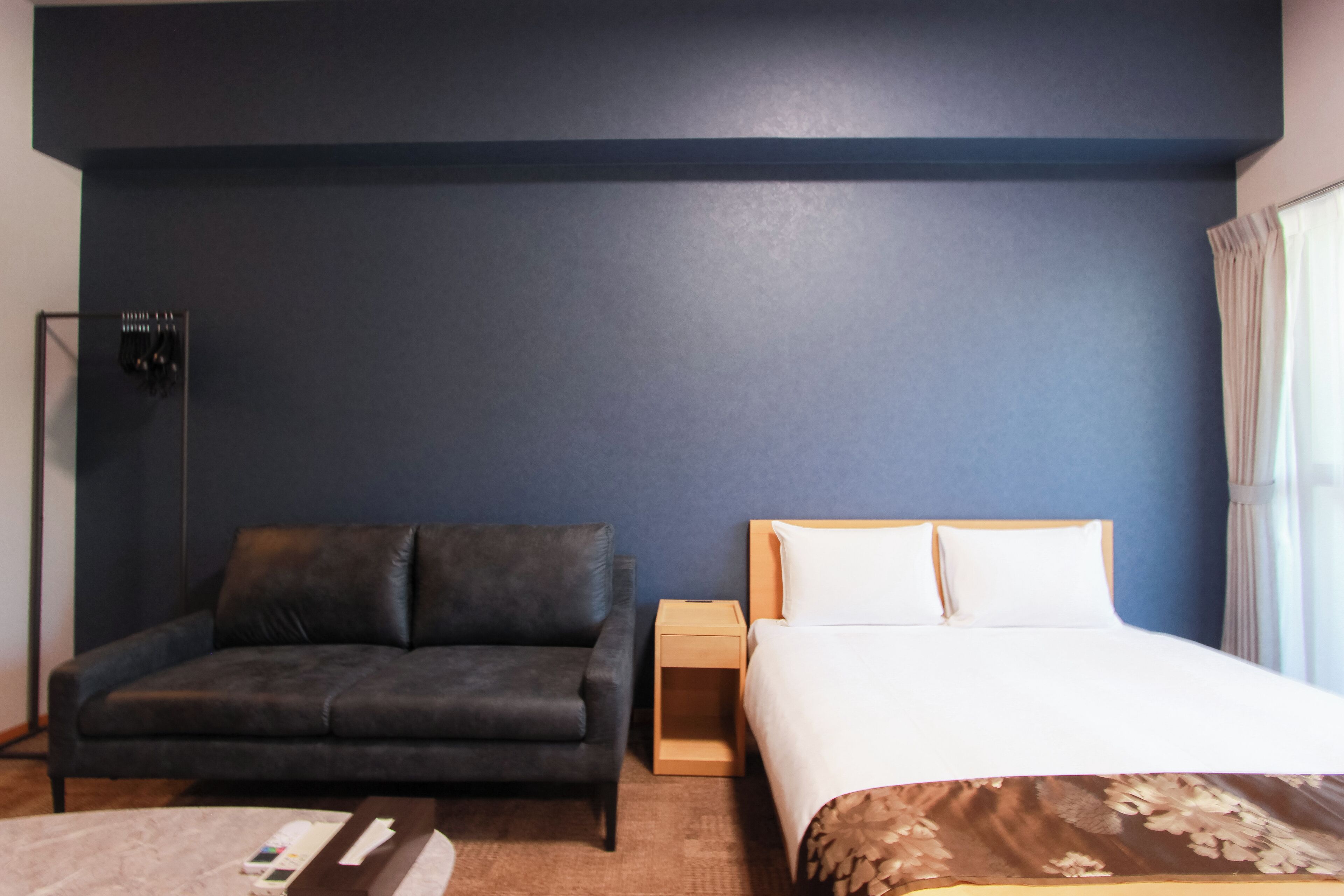 A hotel room with a dark blue wall, a grey sofa, a wooden nightstand, a bed with white linens, and a window with curtains.