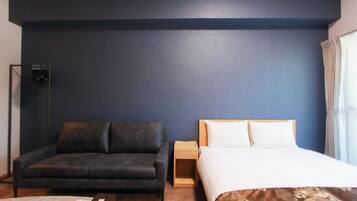 Habitació d'hotel amb una paret blau fosc, un sofà gris, una meseta de nit de fusta, un llit amb roba de llit blanca i una finestra amb cortines.
