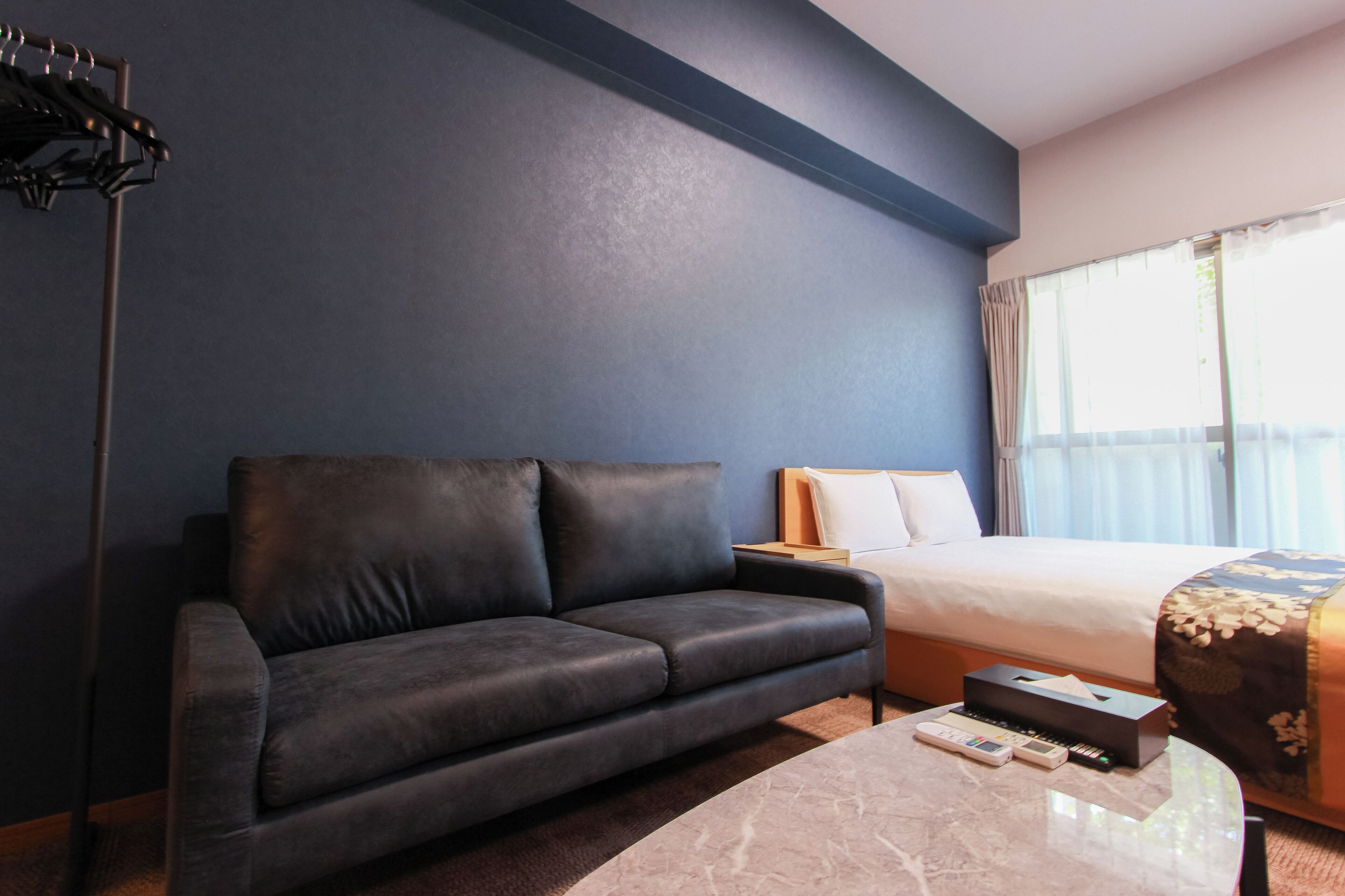Habitación de hotel con pared azul oscuro, un sofá de cuero, cama con colcha estampada, una mesita de noche y una mesa con control remoto y una caja.