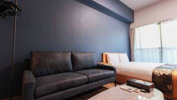 A hotel room with a dark blue wall, a leather sofa, a bed with a floral bedspread, a nightstand, and a table with a remote control and a box.