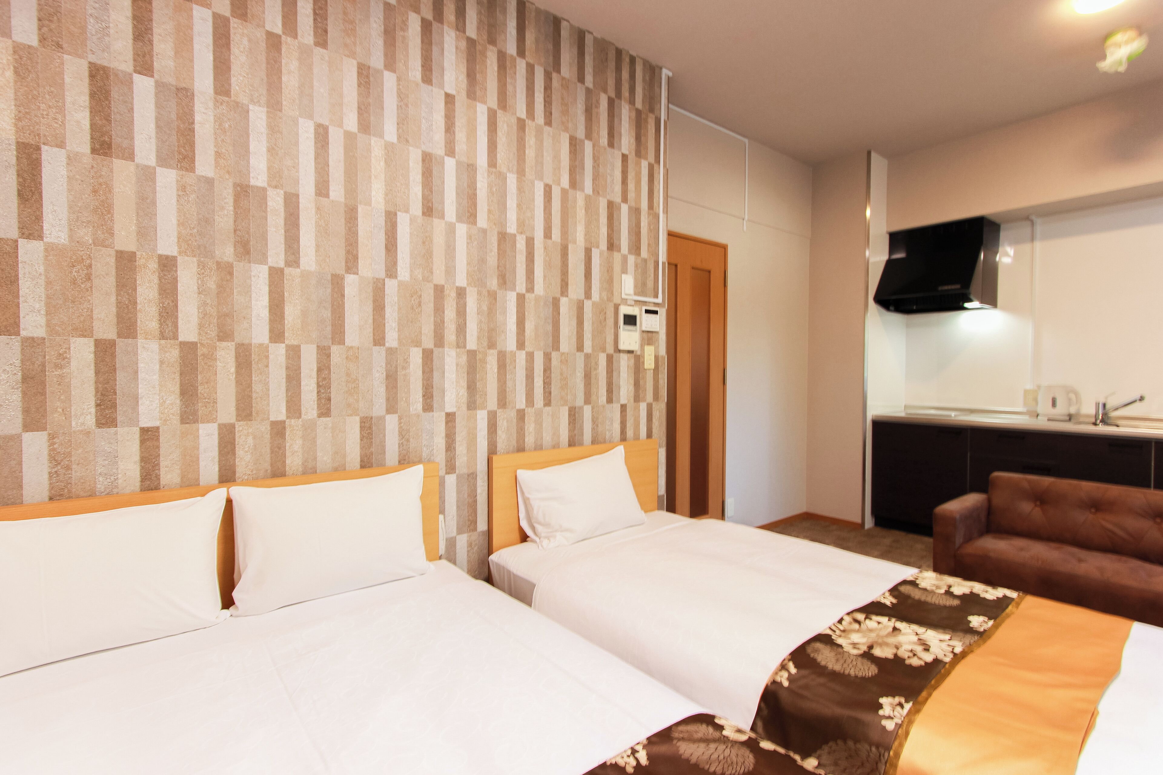 A hotel room with two beds, a wooden headboard, a brown patterned bedspread, a brown leather sofa, a kitchen area with a microwave, and a flat-screen TV mounted on the wall.