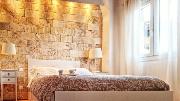 Un dormitori amb paret de pedra, un llit amb roba de llit estampada, dues taules de nit amb làmpades i una finestra amb cortines.