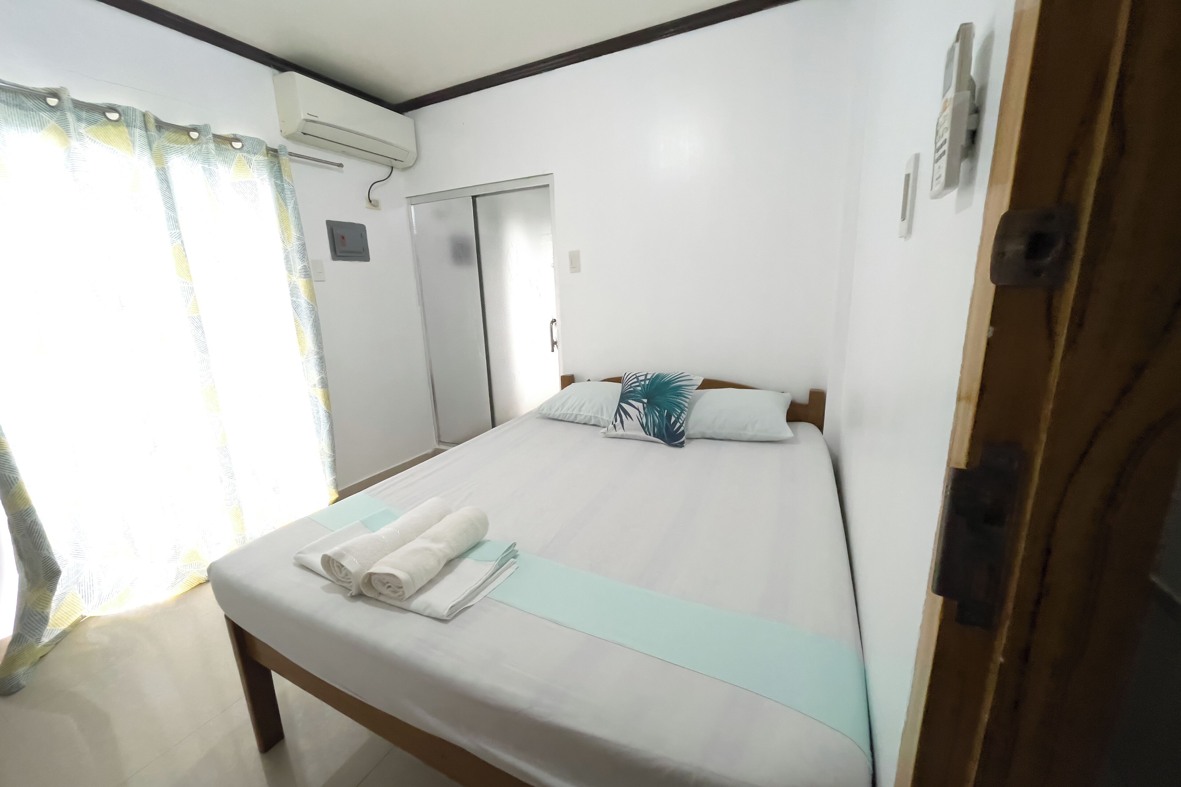 A neatly made bed with a light blue bedspread and two pillows, a wooden nightstand with a folded towel, and a window with curtains.