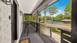 A covered porch with a black railing, a seating area with chairs, and a view of a park with trees and a swimming pool. - The Skipper’s Hideaway: Golf Course & Pool Oasis (Daphne)