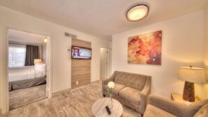 A hotel room with a bed, a sofa, a coffee table with a vase, a TV on the wall, and a floral painting. - Ocean Steps: Pool & Pier Access! (Pompano Beach)