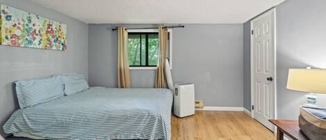 A bedroom with a bed, a window with curtains, a heater, a door, and a lamp on a table.