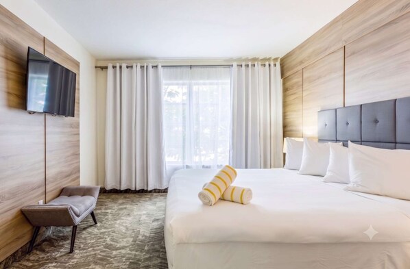 Habitación de hotel con cama, televisor montado en la pared, ventana con cortinas y un pequeño rincón de descanso.