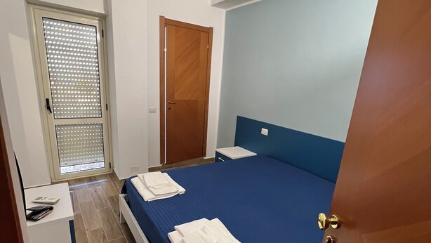 A bedroom with a bed, a desk, a door, and a window with blinds. - B&B CASA LAMANNA- Room No. 5 (Torretta)