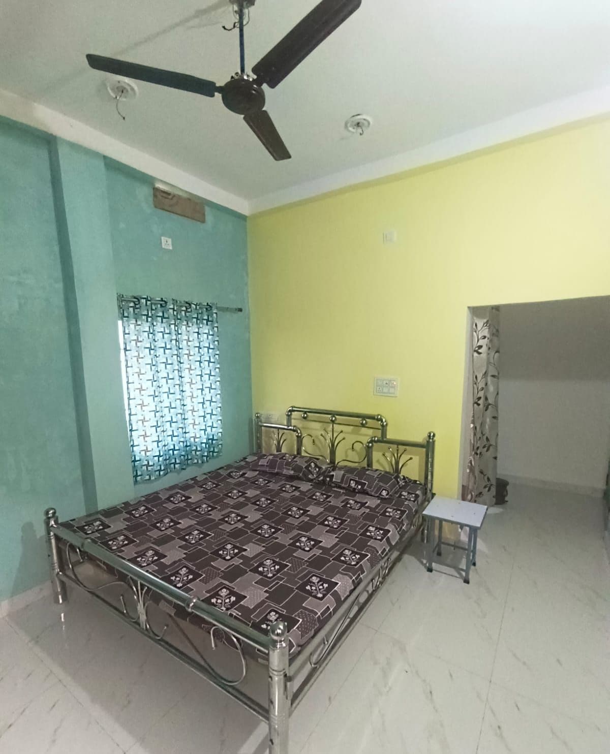 A bedroom with a bed, a ceiling fan, a window with patterned curtains, and a small table.