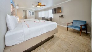 A hotel room with a large bed, a blue chair, a ceiling fan, and a TV on the wall. - Beach Walk Studio w/Pool & 2 Queens (Pompano Beach)