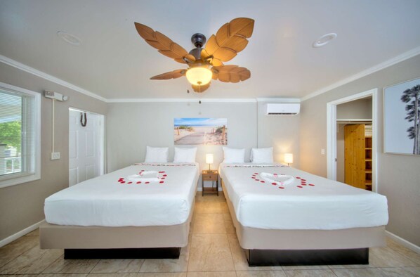 A hotel room with two beds, a ceiling fan, a beach view poster, and a wooden door.