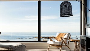 Una habitación moderna con un amplio ventanal con vistas al océano, una silla de madera, una mesita pequeña y una lámpara colgante.