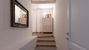 Ein schmaler Flur mit einer Holztreppe, die zu einer Tür führt. Auf der linken Wand hängt ein Spiegel mit einer verzierten Rahmen, der eine Person und ein gerahmtes Bild auf einem Schrank reflektiert.