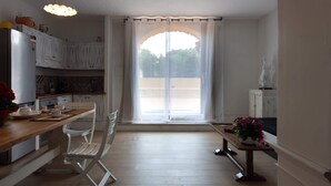 Eine Küche mit weißen Schränken, einem Holzesstisch und Stühlen. Ein Fenster mit durchsichtigen Vorhängen bietet Blick ins Grüne.