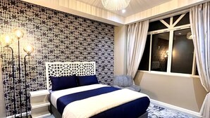 Un dormitorio moderno con un cabecero de estampado geométrico, una cama grande con colcha azul y blanca, un ventanal de techo a piso con cortinas, y una lámpara de techo original.
