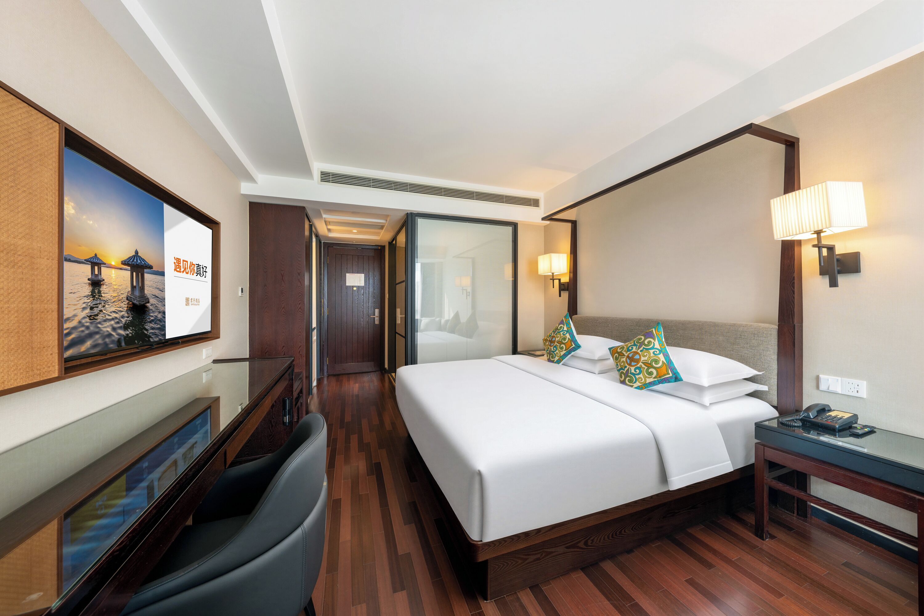 A hotel room with a bed, bedside table, lamp, and a view of a corridor.