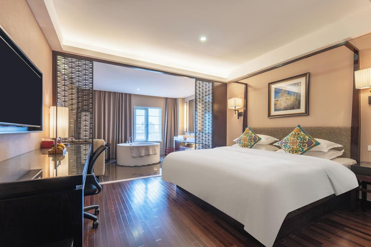 Ein Hotelzimmer mit einem großen Bett, einem Schreibtisch mit Stuhl, einem Fernseher, einem Badezimmer mit Badewanne und einem Fenster mit Vorhängen.
