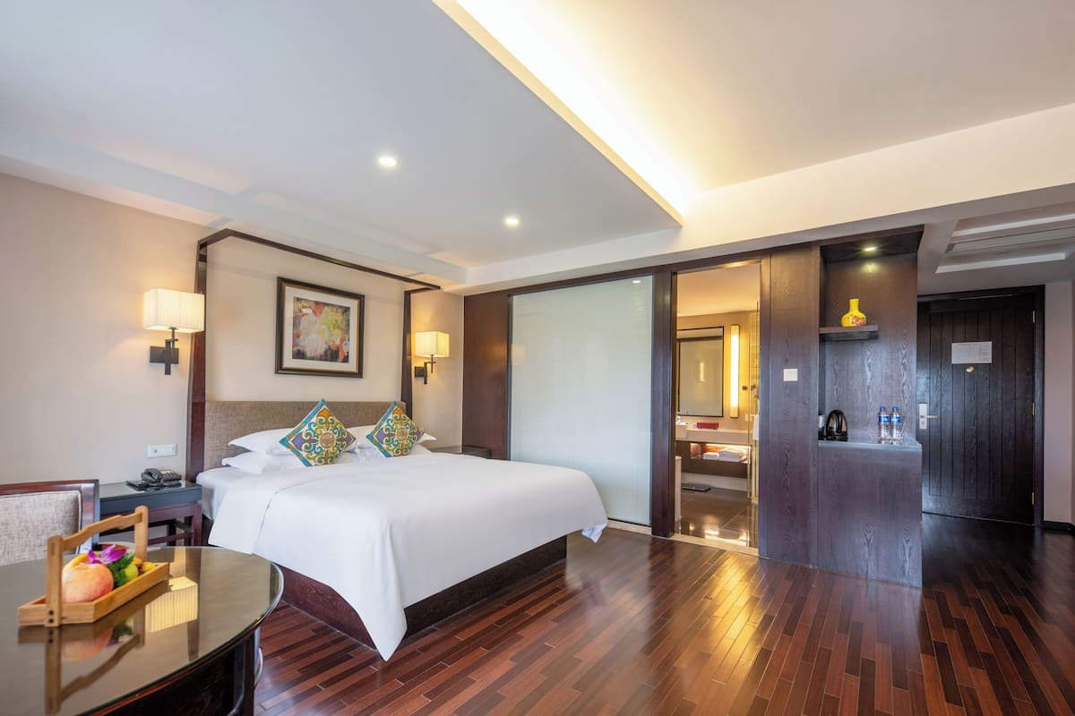 Ein Hotelzimmer mit einem großen Bett, Nachttischlampen, einem Holztisch und einem kleinen Tisch mit einer Obstauswahl.