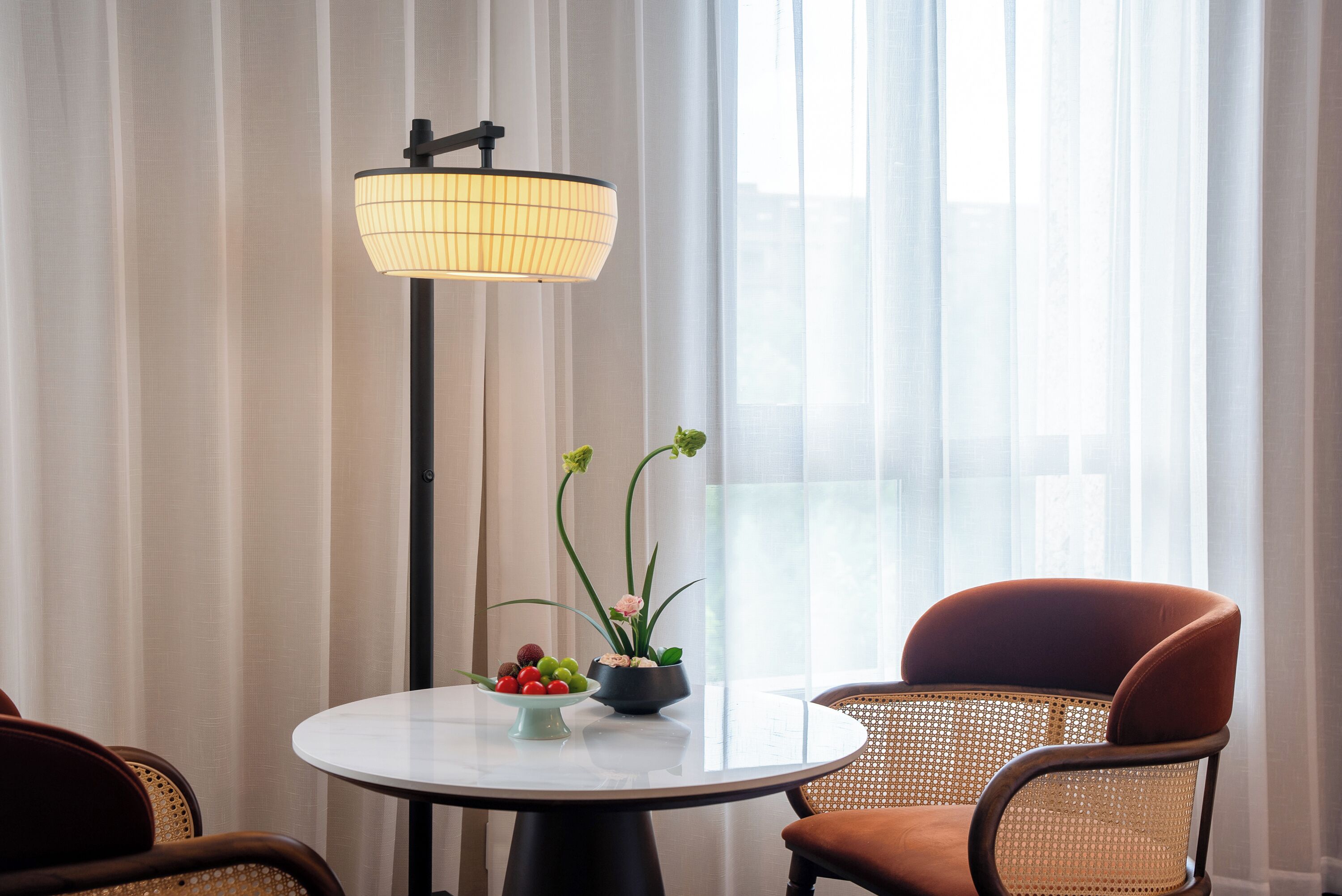Una habitación con una mesa redonda, dos sillas, una lámpara de pie y un ventanal con cortinas translúcidas.