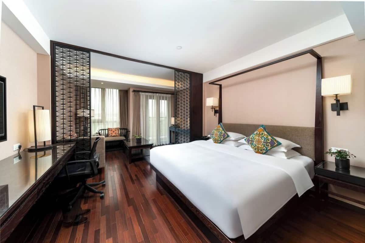Ein Hotelzimmer mit einem großen Bett, einem Schreibtisch und einem Stuhl. Es gibt ein Fenster mit Vorhängen, eine Wandlampe und einen dekorativen Paravan.