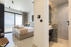 A modern hotel room with a large bed, a bathroom with a glass shower, and a view of the city. - Bella Casa - Newly Built Designer Villa (Kalyves)