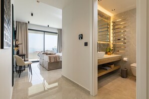 A modern hotel room with a large bed, a sitting area with a chair, a view of the sea, and a bathroom with a marble countertop and towel rack. - Bella Casa - Newly Built Designer Villa (Kalyves)