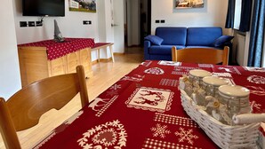 Ein Wohnzimmer mit Fernseher, einer Couch, einem Esstisch mit rotem Tischläufer und einer kleinen Küchenzeile mit Holzschrank.