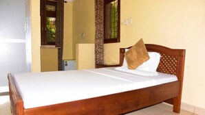 Una cama individual con estructura de madera y ropa de cama blanca, una mesita de noche pequeña y una ventana con barrotes.