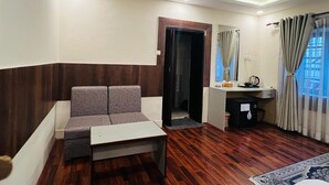 Una sala de estar moderna con un sofá gris, una mesita blanca y piso de madera. Hay una puerta que da a un baño, y una ventana con cortinas.