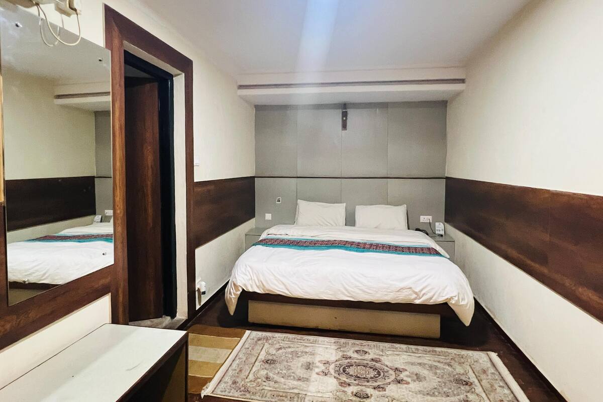 A hotel room with a bed, a bedside table, a mirror, and a patterned rug.