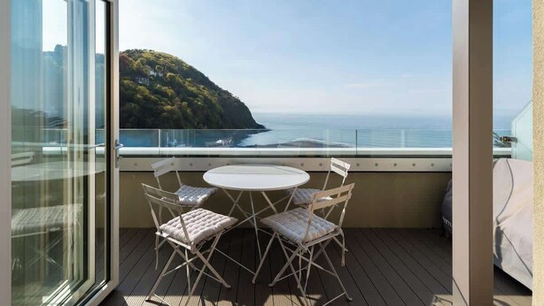 A balcony with a table and chairs, overlooking a coastal landscape with a hill and the sea. - Oceanfront Apartment w/Balcony & Sauna, Pet-Friendly (Lynmouth)