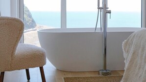 A modern bathroom with a freestanding bathtub, a chair, and a view of the ocean. - Oceanfront Apartment w/Balcony & Sauna, Pet-Friendly (Lynmouth)