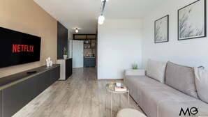 Ein modernes Wohnzimmer mit einem Flachbildfernseher, der das Netflix-Logo zeigt, einem grauen Sofa, einem kleinen runden Tisch mit einem Buch und einer Glaseinmachglas und einer Küche im Hintergrund.