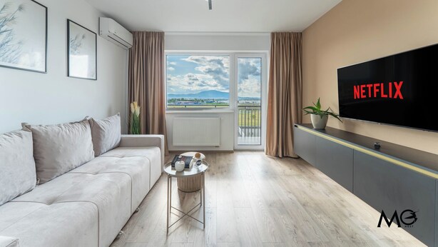 Ein modernes Wohnzimmer mit einer Couch, einem Couchtisch, einem an der Wand montierten Fernseher und einem großen Fenster mit Blick ins Freie.
