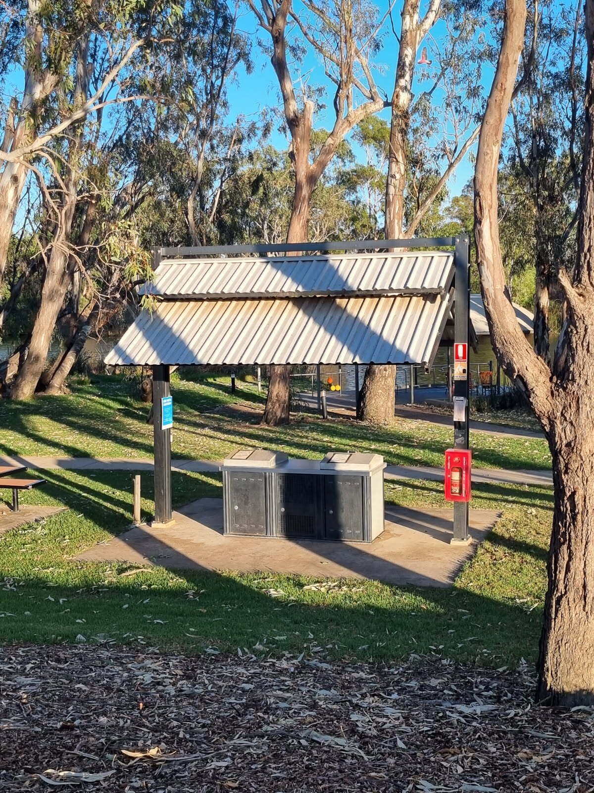 Un refugio en el parque con techo metálico, bancos y un cubo de basura.