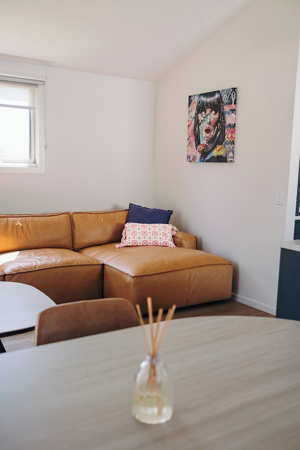 A living room with a brown leather sofa, a small table with a glass vase, and a colorful abstract painting on the wall.