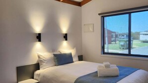 A bedroom with a large bed, two bedside tables, a window with a view, and a sloped ceiling with wooden beams. - Bridgewater Caravan Park (Bridgewater on Loddon)