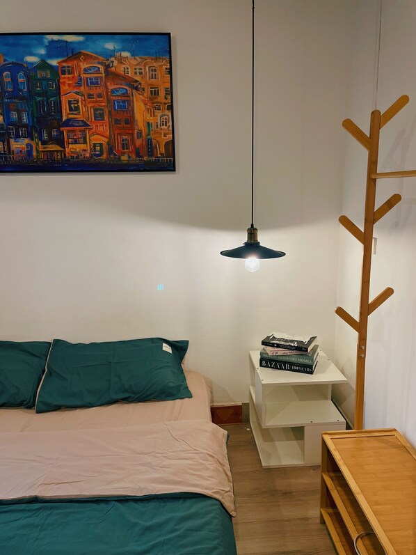 A bedroom with a bed, a hanging light, a wooden coat rack, a small white shelf with books, and a painting on the wall.