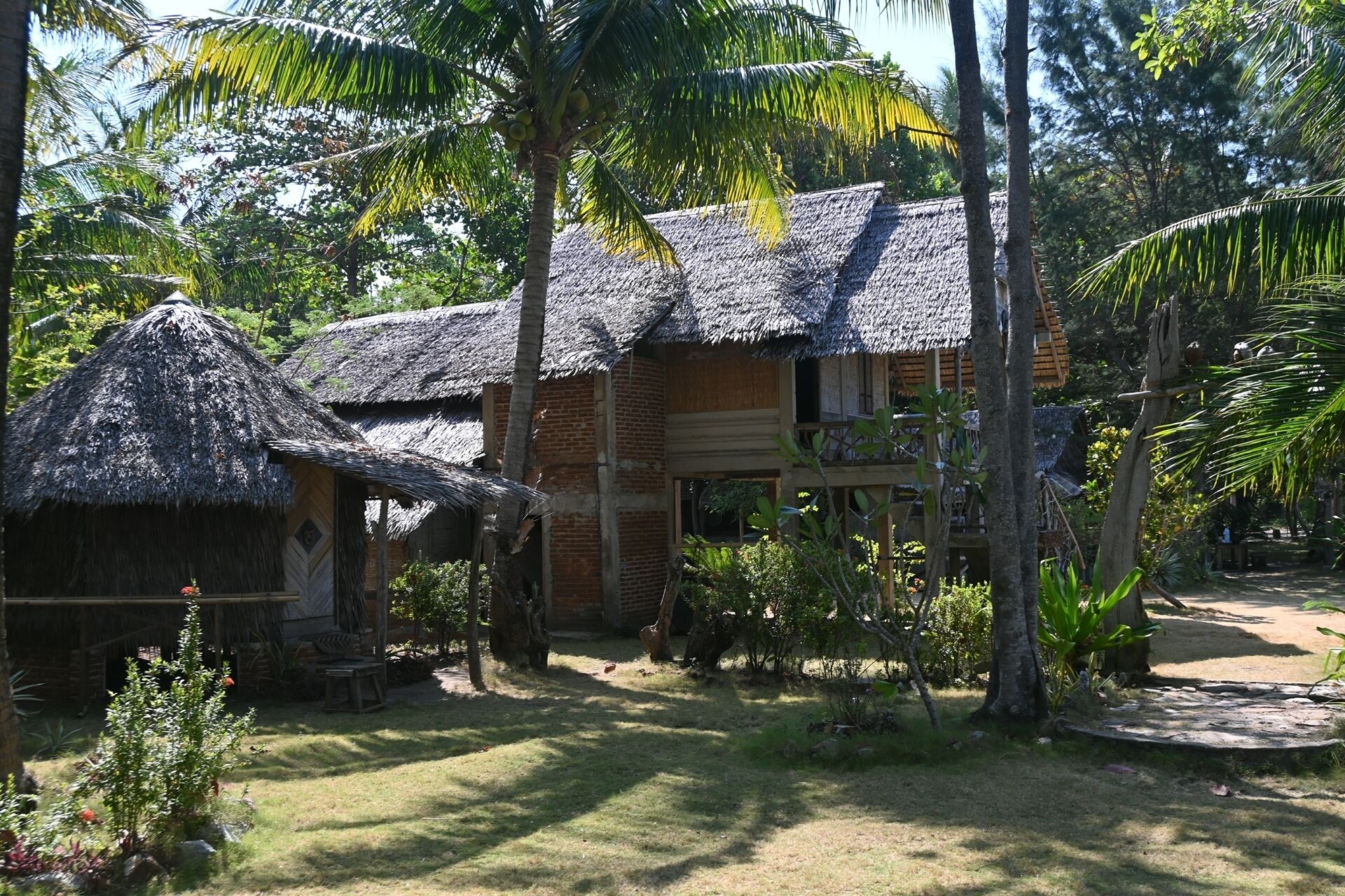 A traditional thatched-roof house with a wooden balcony, surrounded by lush greenery and palm trees.