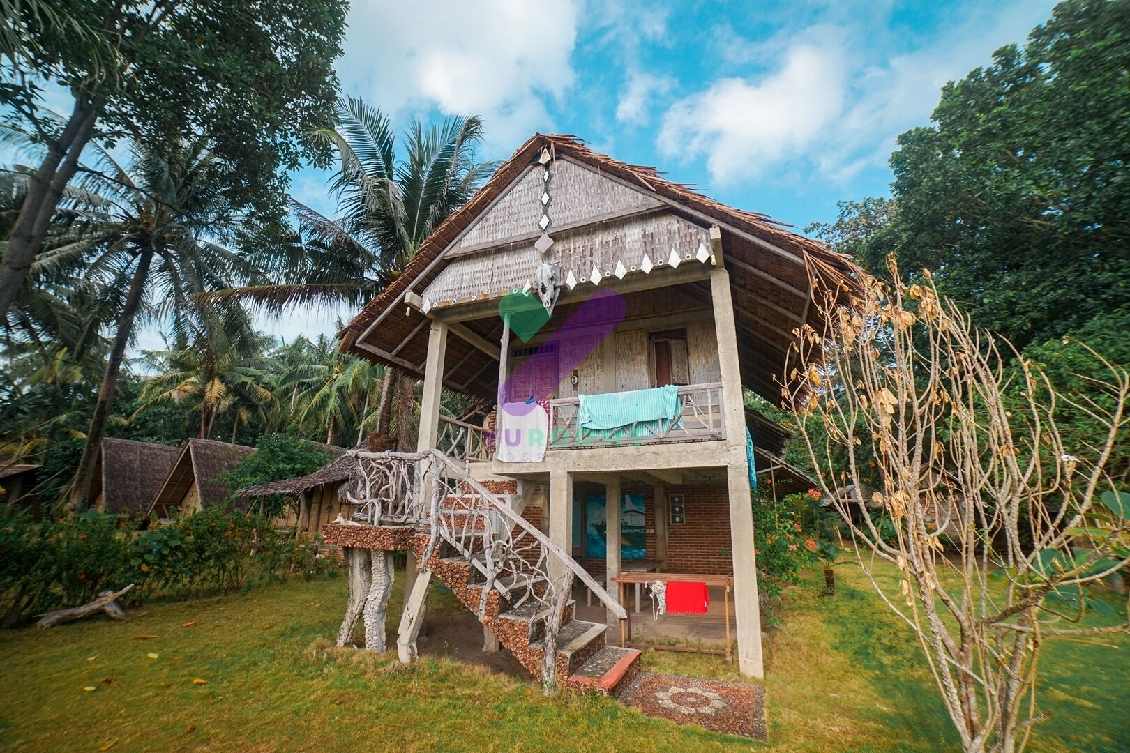A two-story wooden house with a balcony, surrounded by tropical vegetation.