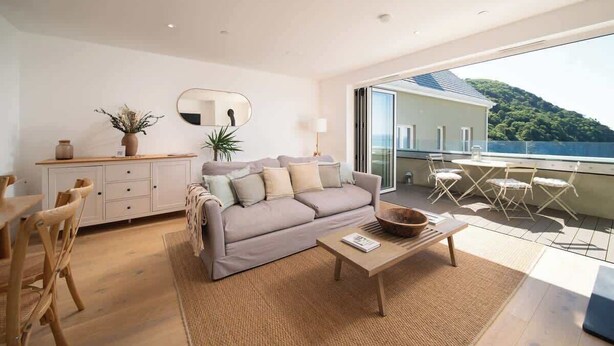 A modern living room with a sofa, coffee table, and dining area. There is a balcony with a view of a hill. - Panoramic Sea Views, Private Balcony & Sauna (Lynmouth)