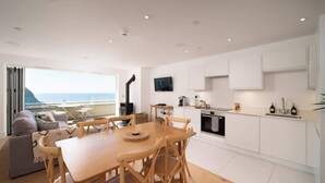 A modern kitchen with white cabinets, a dining table with wooden chairs, and a TV mounted above the counter. - Panoramic Sea Views, Private Balcony & Sauna (Lynmouth)