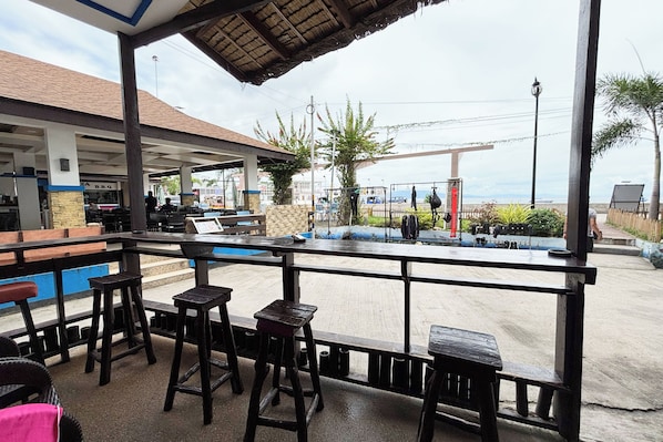 An outdoor seating area with wooden stools and tables, a covered roof, and a view of a gas station and beach in the background.