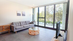 A modern living room with a grey sofa, a round coffee table, and a flat-screen TV. - Olympic Park Stylish 2BR | Walk to stadium | Pool & Gym (Lidcombe)