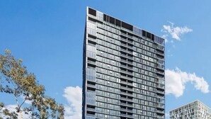 A modern high-rise building with a grid-like facade, multiple balconies, and a mix of glass and concrete materials. - Olympic Park Stylish 2BR | Walk to stadium | Pool & Gym (Lidcombe)