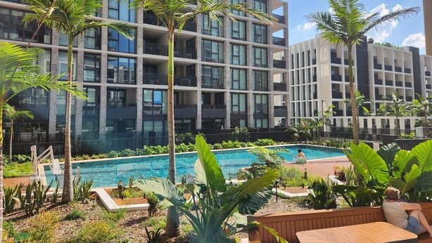 A modern hotel with a swimming pool surrounded by palm trees and greenery. - Olympic Park Stylish 2BR | Walk to stadium | Pool & Gym (Lidcombe)