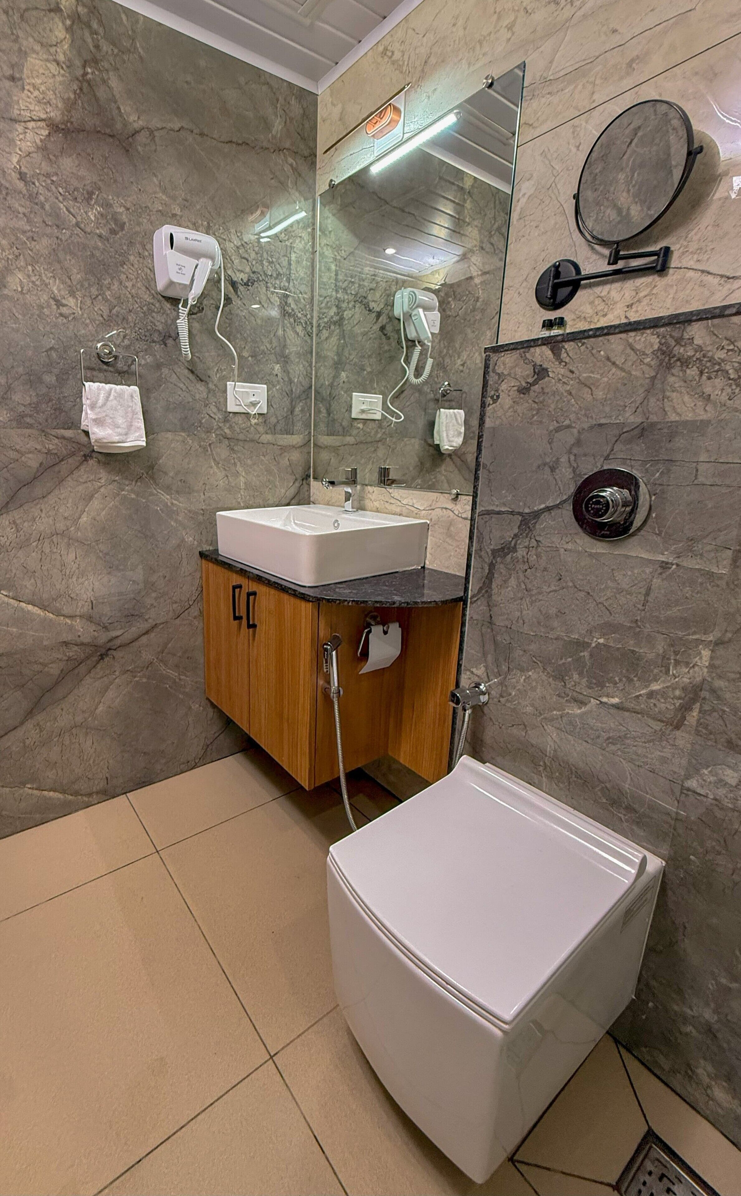 Une salle de bain moderne avec un mur en marbre, un meuble en bois, un lavabo blanc, un miroir, un sèche-cheveux et des toilettes.