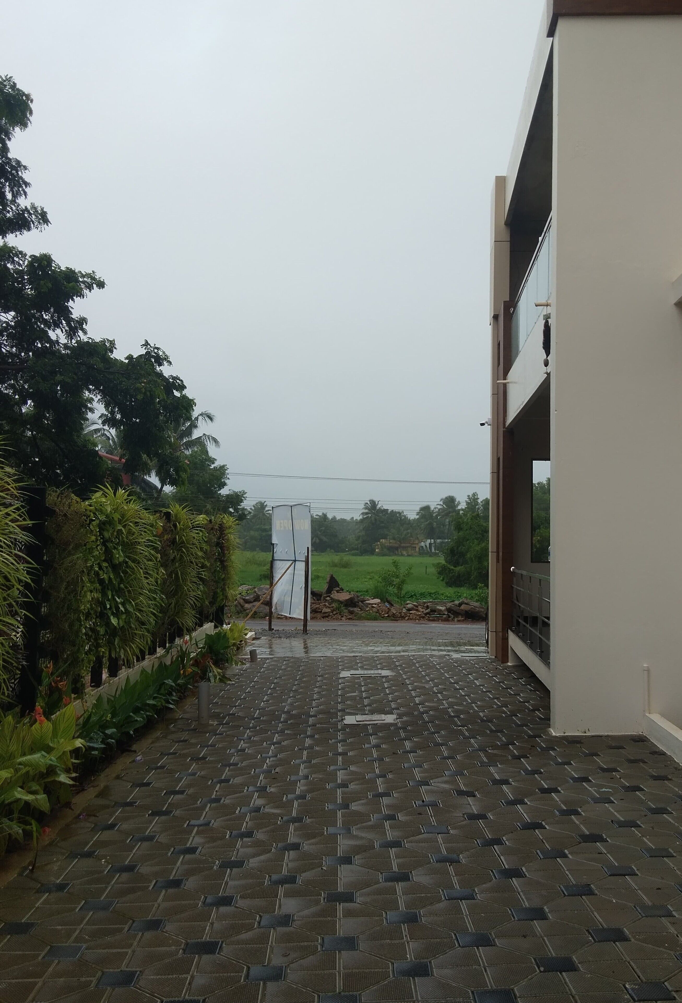 Um caminho pavimentado leva a um acesso de edifício, cercado por vegetação e com vista para um campo.