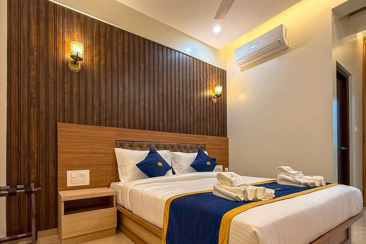 A modern hotel room with a wooden headboard, a bed with blue and white bedding, a nightstand, a bedside table, a ceiling fan, and an air conditioning unit.