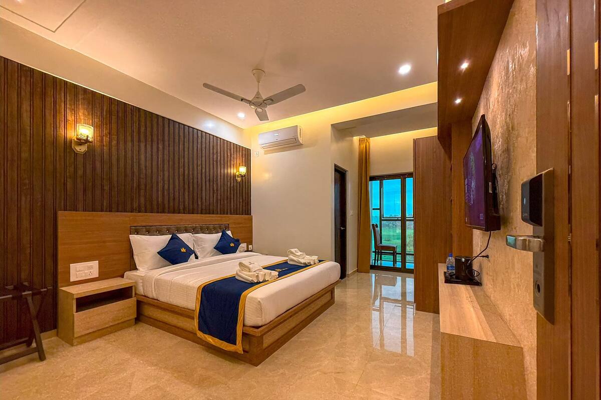 A modern hotel room with a wooden headboard, a flat-screen TV, a ceiling fan, and a view of the outdoors through a large window.